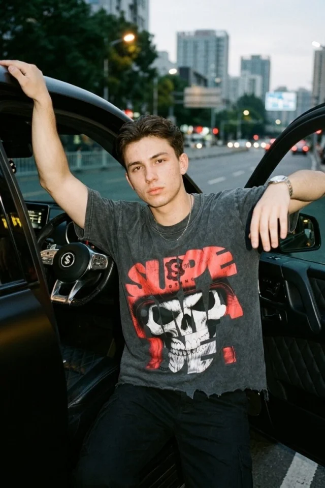 A young man in the washed skull-graphic tee leaning against a car door on a city street at dusk.