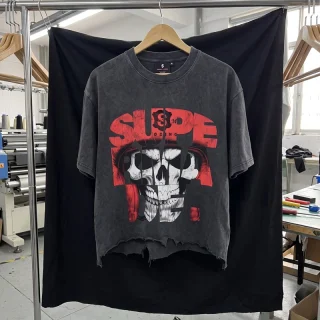 A washed charcoal T-shirt with a red and white skull graphic and a frayed hem, on a hanger in front of a black cloth in a workshop.