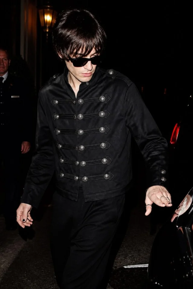 A model in dark glasses wearing a black leather jacket with rows of silver buttons across the chest, on a city street at night.