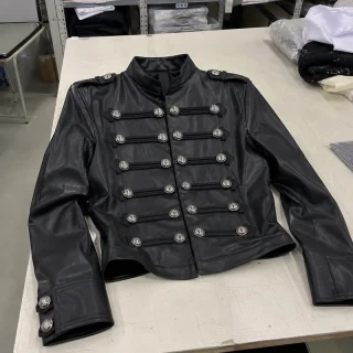 A black leather jacket with silver buttons and braid across the front, laid on a white worktable.