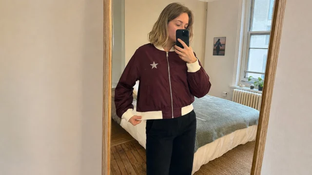 A mirror selfie on a phone of someone in a burgundy bomber jacket with a cream collar and cuffs.