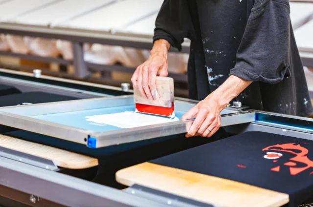 A printer pulling ink through a screen onto a black T-shirt on a print table.