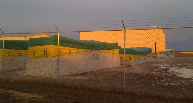 Large cotton modules stored beside a warehouse and cotton gin before processing.