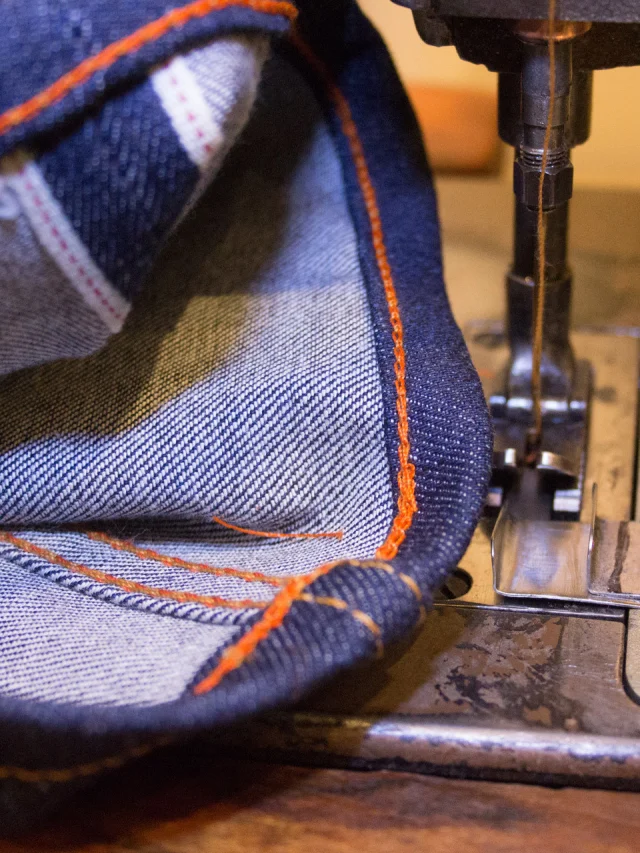 A denim hem being sewn with orange thread under the needle of a sewing machine.