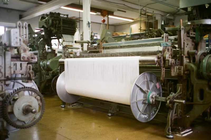 An industrial loom winding white woven cloth onto a roll.