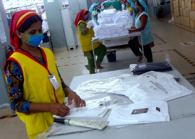A worker in a factory packing room putting folded white T-shirts into clear polybags.