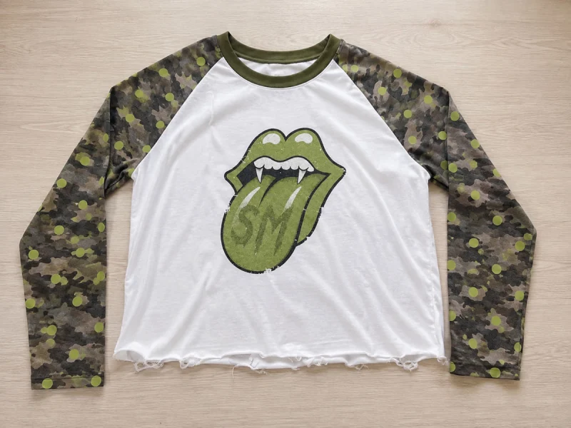 A cropped white raglan top laid flat on a wooden floor. It has camouflage sleeves with lime-green dots, an olive rib neckline, a raw hem and a large green front graphic.