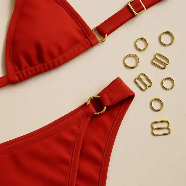 A red triangle bikini top and bottom joined with gold rings, with loose gold rings and strap sliders on a cream surface.