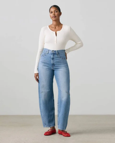 A woman in blue barrel jeans, a cream top and red flats.