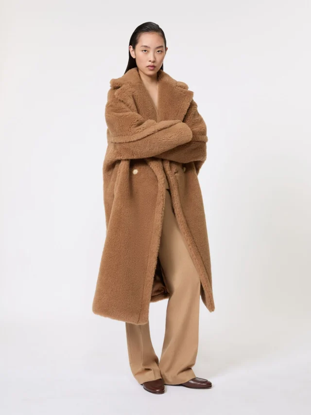 A camel teddy coat with large lapels and wide sleeves, worn over beige trousers.