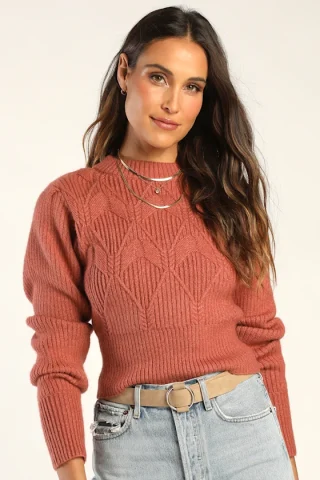 A woman in a rust cropped sweater with cable and openwork panels and a high ribbed hem.
