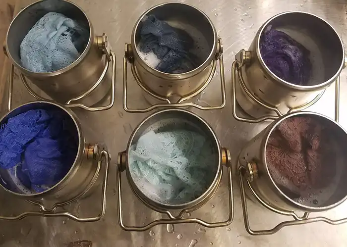 Dyed fabric samples in small steel canisters on a lab bench, in blues, purples and a pale green.