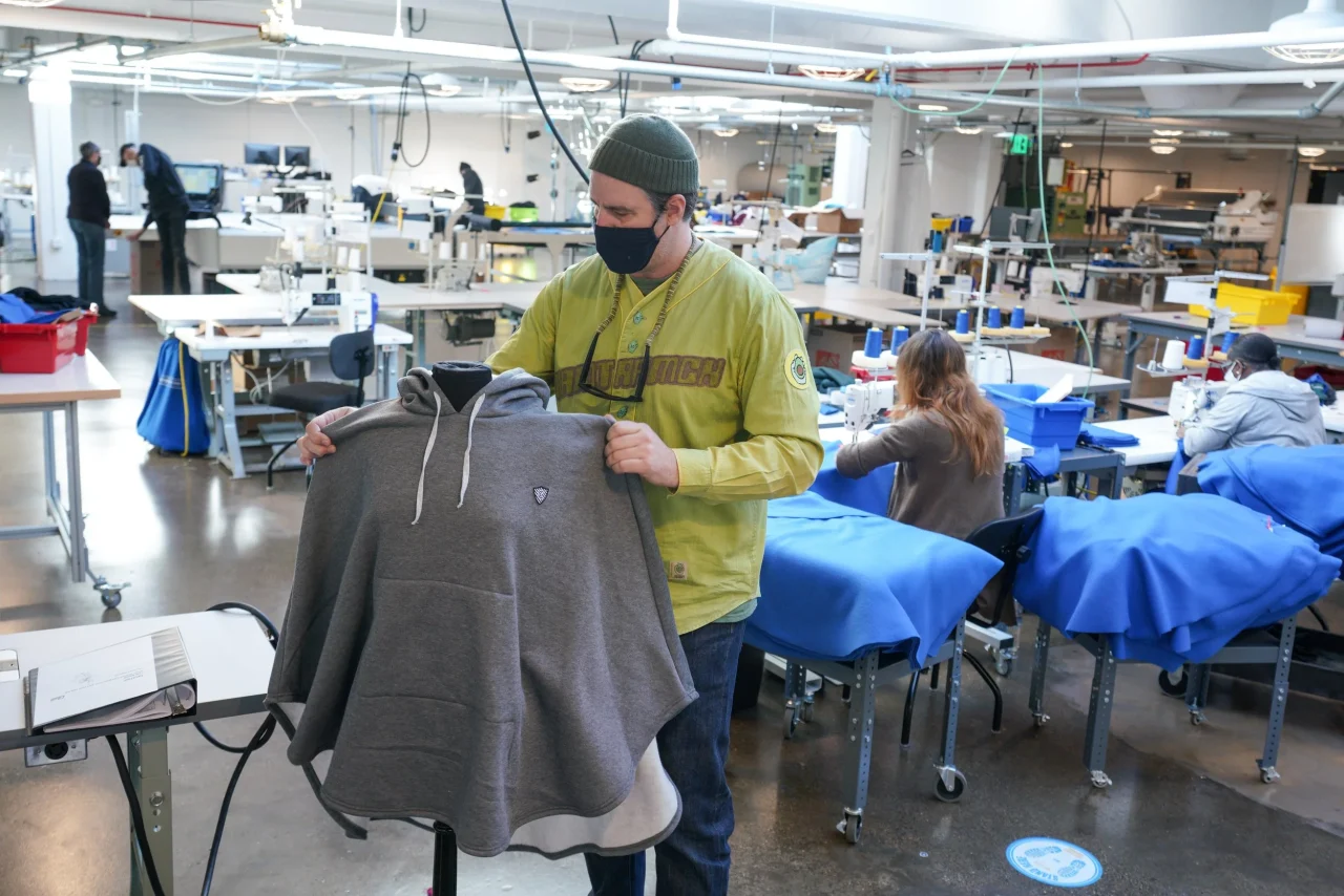 An inspector holding up a gray hoodie on a factory floor with sewing tables behind him.