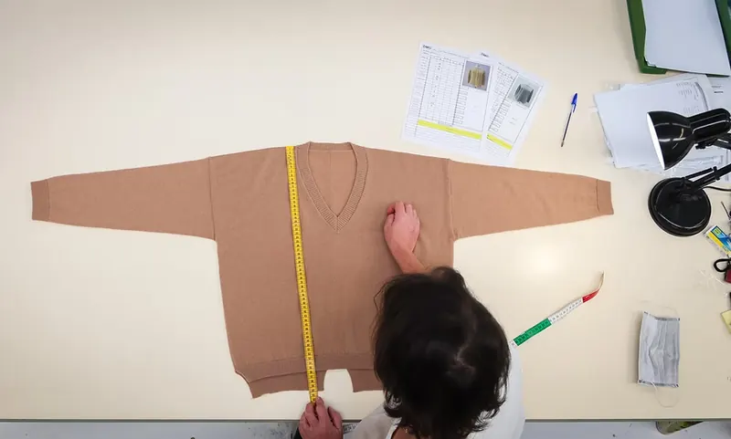 An inspector measuring a tan knitted sweater laid flat on a worktable.