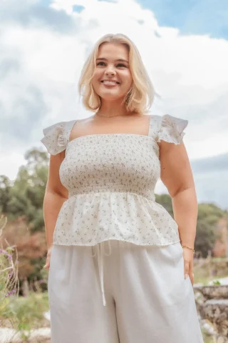 A floral blouse with a shirred bodice, ruffled shoulder straps and a small peplum, worn over cream trousers.