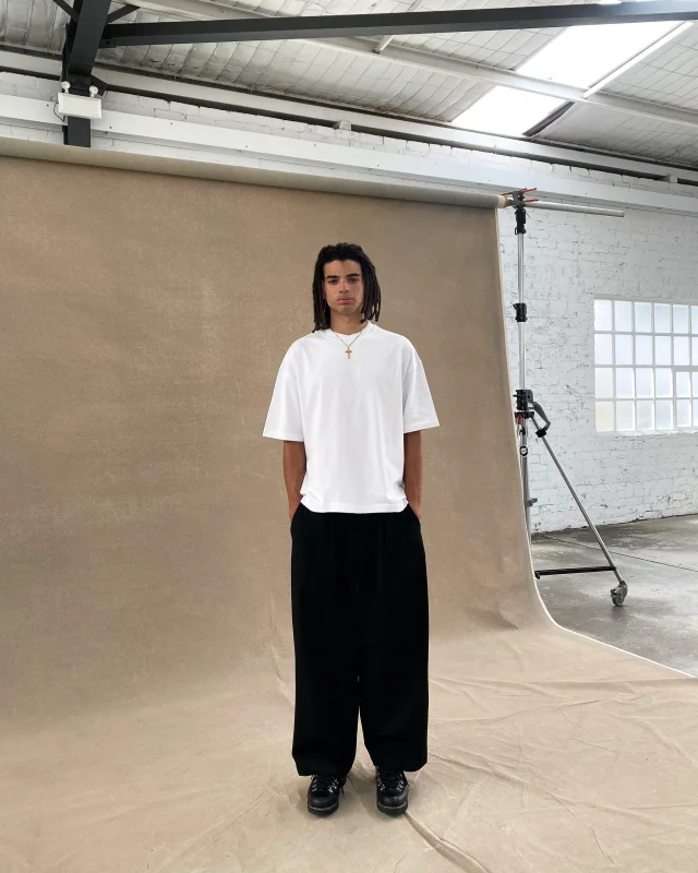A man in an oversized white T-shirt and wide black trousers in a photo studio.