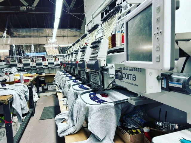 Gray hoodies hooped on a row of multi-head embroidery machines in a workshop.