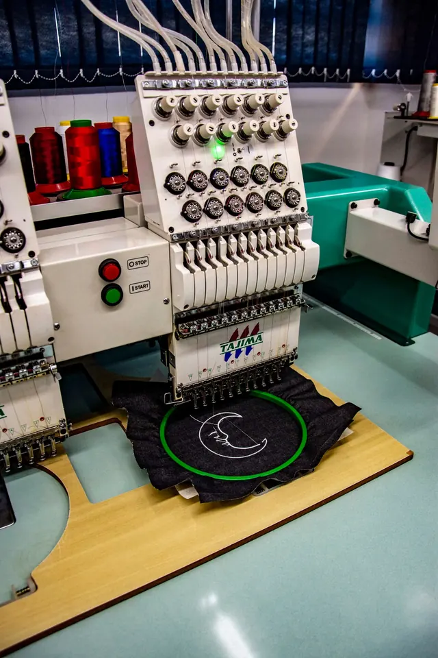 A multi-head embroidery machine stitching a design onto a black garment panel.