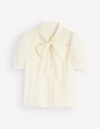 An ivory silk blouse laid flat, with a large bow tied at the neck, short gathered sleeves and a button front.
