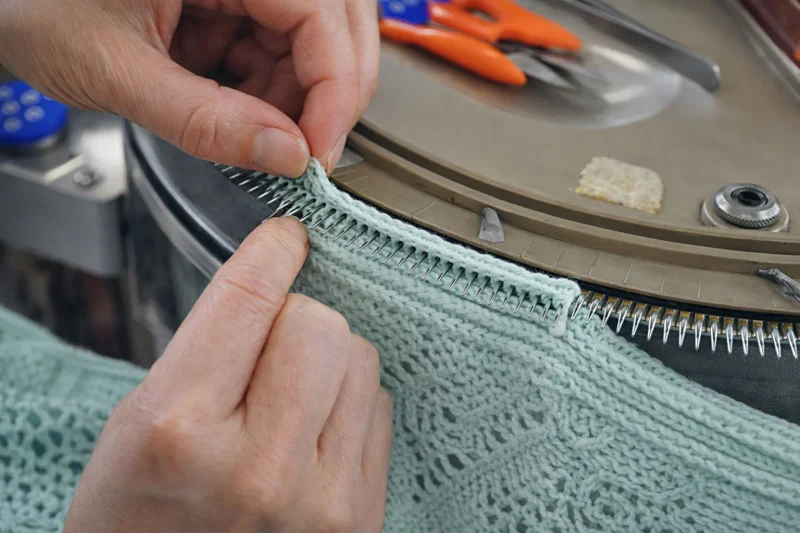 Hands feeding the edge of a mint-green knit panel onto a linking machine.