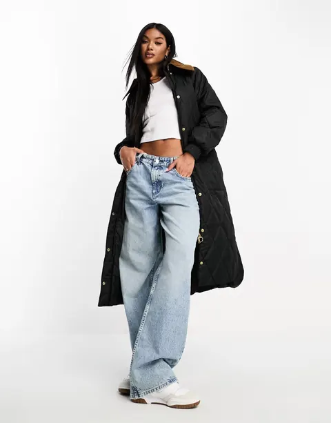 A model in a long black quilted coat worn open over a white top and wide light jeans.