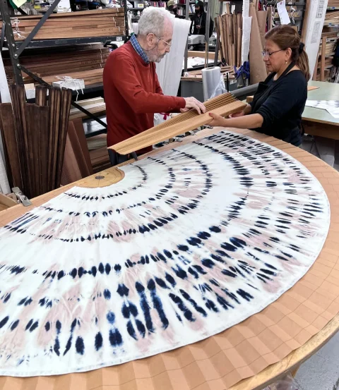Two pleaters lining up a round printed fabric panel on a long pleating table.