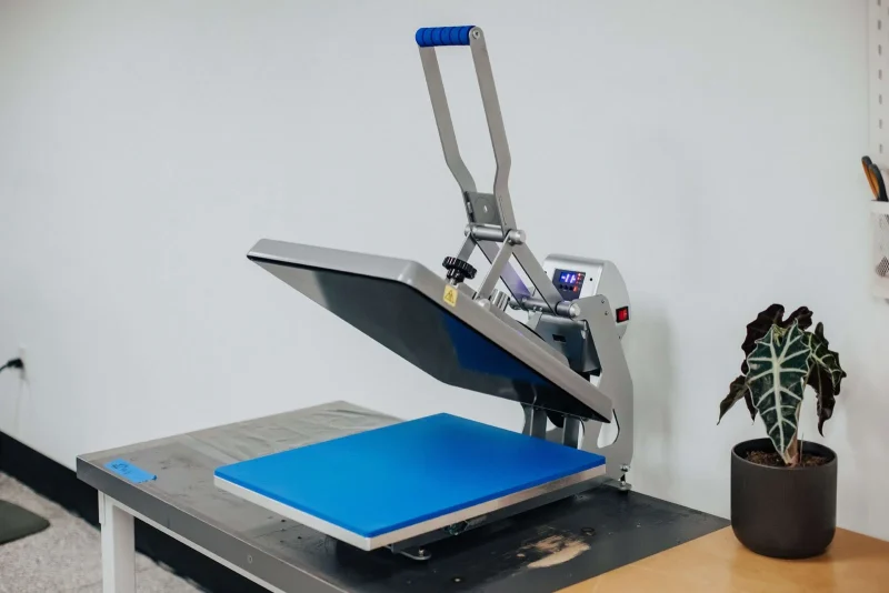 A clamshell heat press with a blue pad on a workbench beside a potted plant.