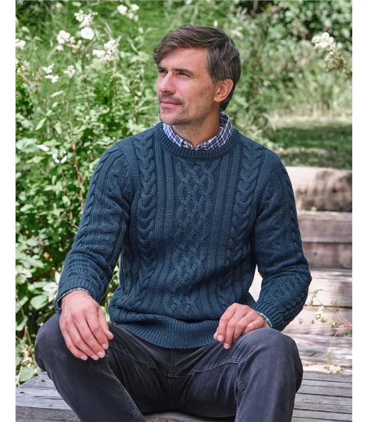 A man in a navy cable-knit sweater sitting outdoors in a garden.