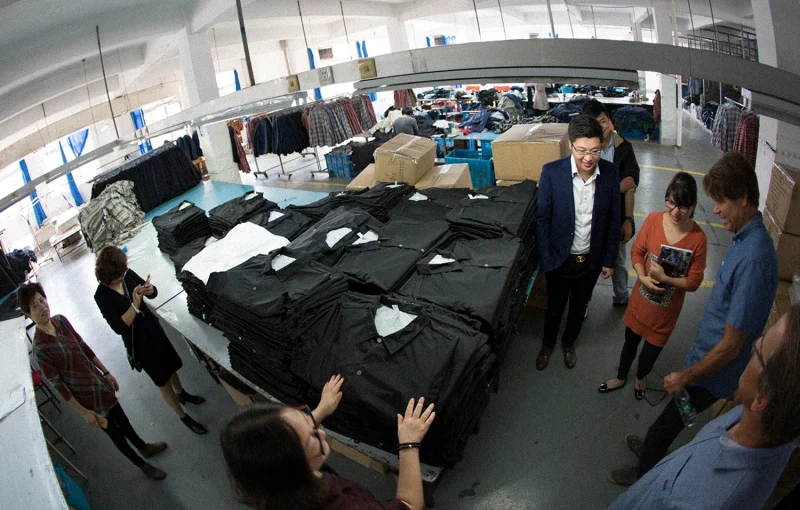 Visitors looking over stacks of black garments on tables inside a clothing factory.