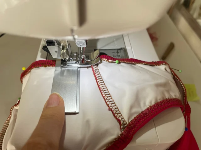 A white swimsuit with red zigzag-stitched edges being sewn at a sewing machine, held flat with pins.