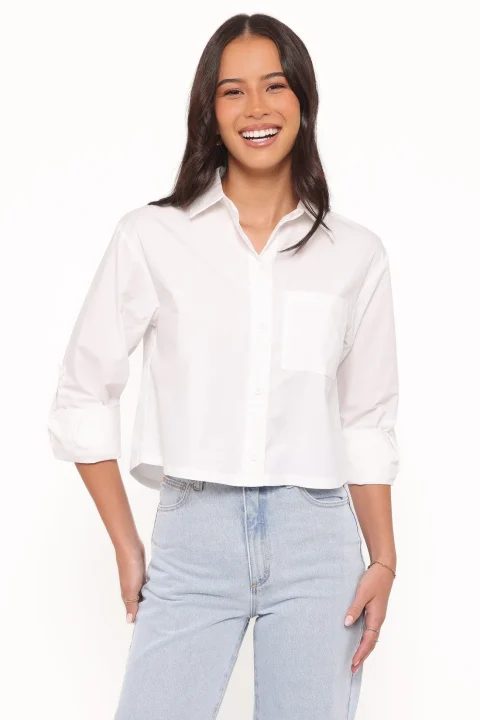 A white cropped button-front blouse with a boxy body and rolled sleeves, worn over light blue jeans.