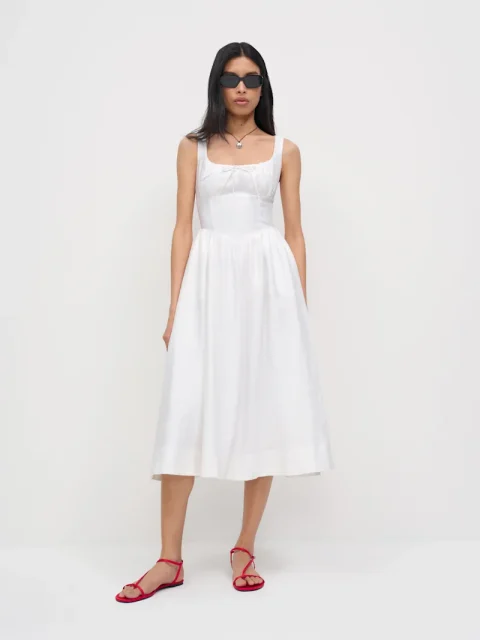 A woman in a white linen sundress with shoulder straps and a full skirt, worn with red flat sandals.