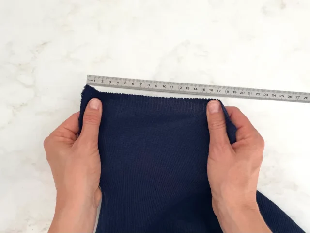 Two hands stretching navy jersey along a ruler to measure how far it gives.
