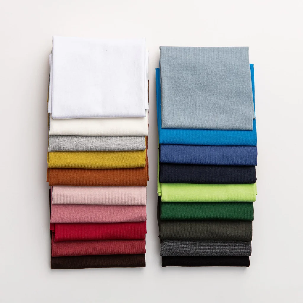Folded cotton jersey fabrics stacked in white, gray, red, yellow, pink, blue, and green.