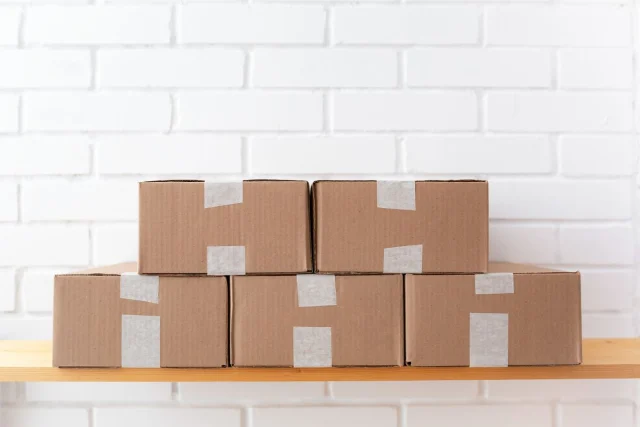 Five sealed cardboard shipping cartons stacked on a wooden shelf against a white brick wall.