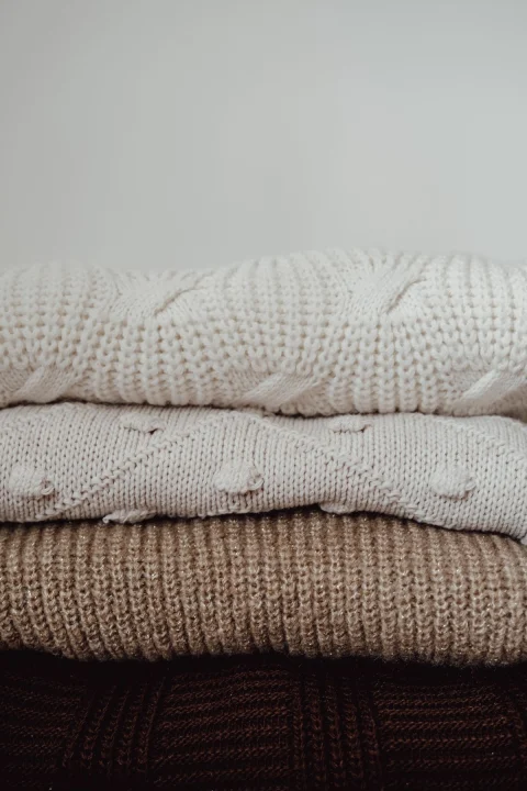 A stack of folded knit sweaters in white, cream, tan and dark brown on a light background.