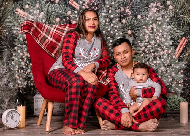 Parents in matching gray reindeer-print tops and red buffalo-plaid pajama pants, holding a baby, in front of Christmas trees.