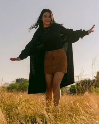 Woman in a long black open cardigan over a short brown skirt in a grass field.