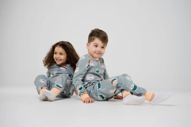 Two children in matching snug-fit printed pajamas sitting on a white floor.