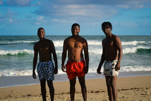 Three men on a beach in short swim trunks, one in a red solid pair and two in printed pairs.