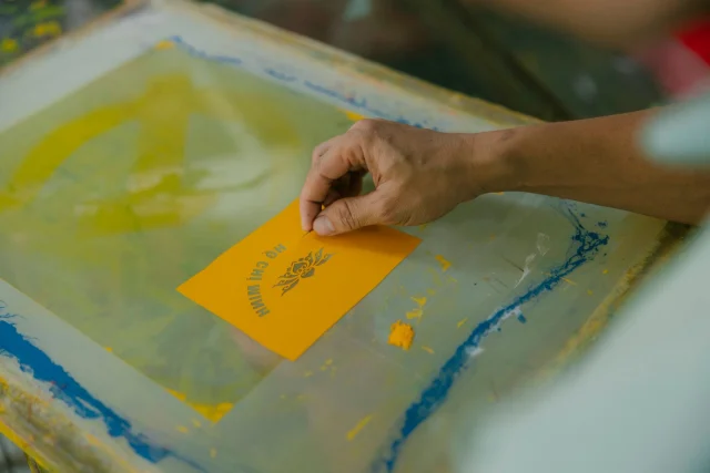A hand lifting a freshly printed yellow sheet off a screen.