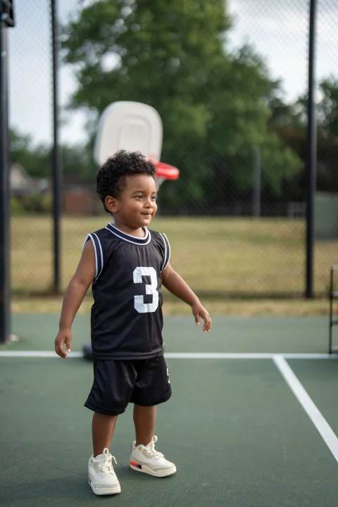 A toddler in a black basketball jersey with the number 3 and black shorts on an outdoor court.