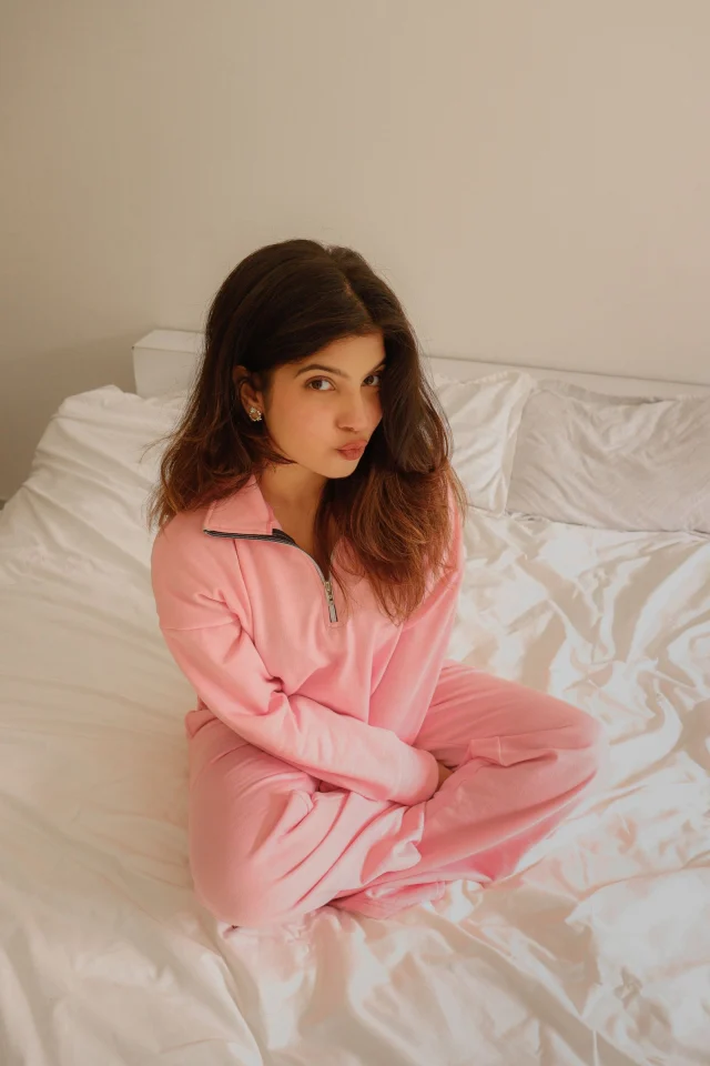 A woman in a pink lounge set with a zip collar sitting on a bed.