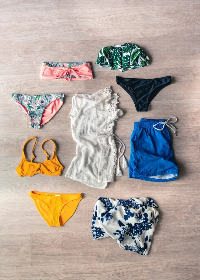 Bikini tops and bottoms, swim trunks and a white cover-up laid out on a wooden floor.