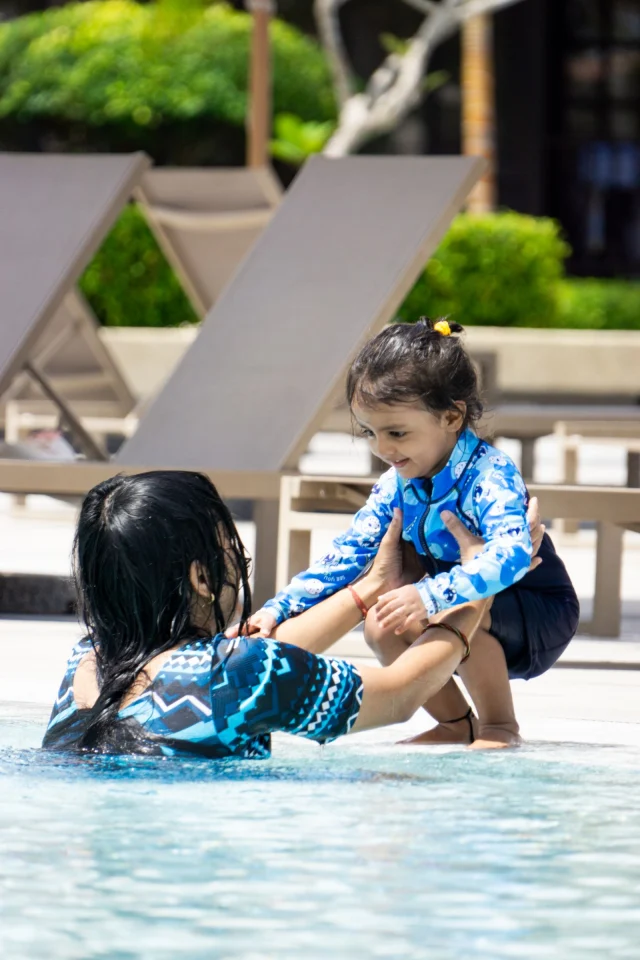 A toddler in a long-sleeve zip-front rash guard reaching for a woman in a resort pool.