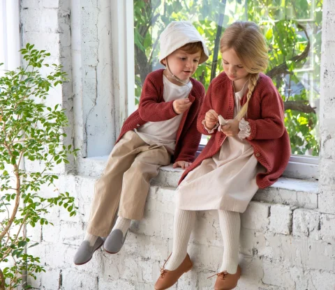 Two children in red knitted cardigans sitting on a white brick windowsill.