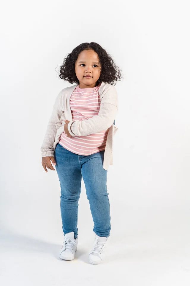 Little girl wearing a cream cardigan over a pink striped top and jeans.