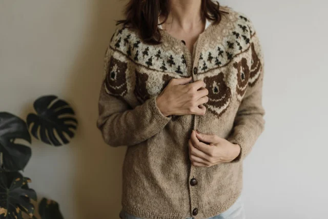 A woman fastening the buttons on a beige cardigan with a patterned yoke.