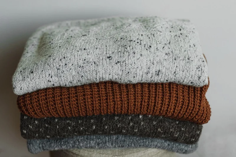 Folded knit sweaters in grey, rust and charcoal stacked on a table.