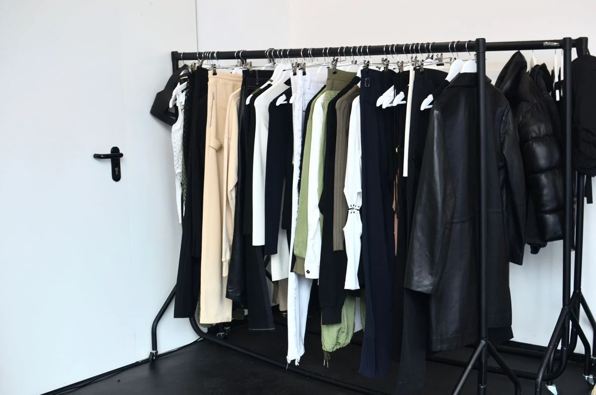 A clothes rail in a white showroom with trousers, shirts, a leather-look coat and a puffer jacket on hangers.
